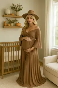 A maternity fashion model with light blonde hair wearing an elegant mocha-toned gown stands beside a warm wood crib in a sunlit Mocha Mousse nursery decorated with ornamental pumpkins, gourds, and vases filled with beauty berries and greenery.
