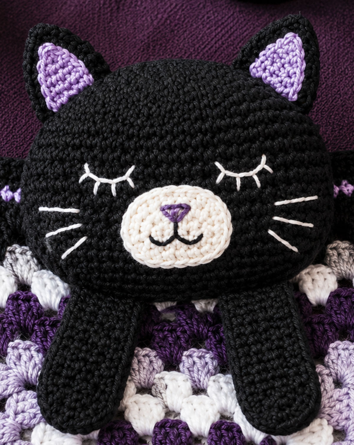 Close view of a black cat crochet lovey with light embroidered eyes, contrast muzzle, pointed ears, and short whiskers