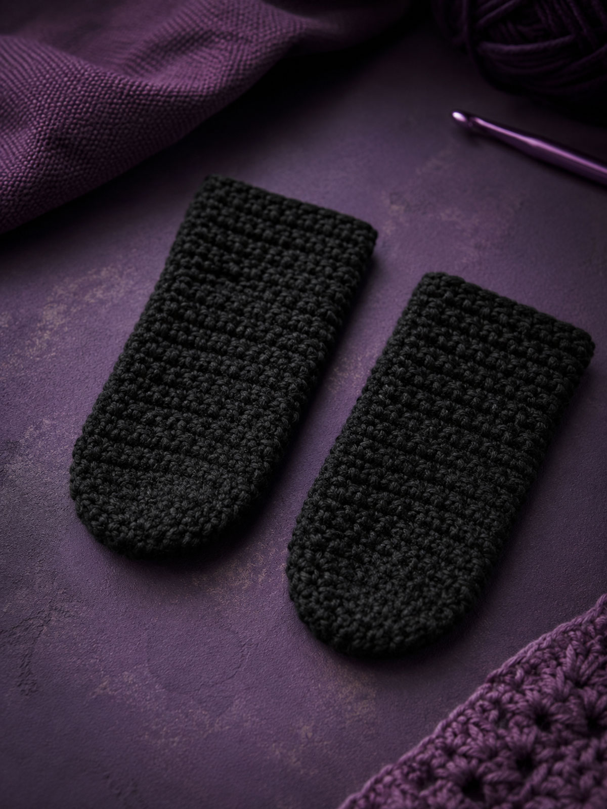 Two flat unstuffed black crochet cat paws arranged side by side on a dark purple work surface before assembly