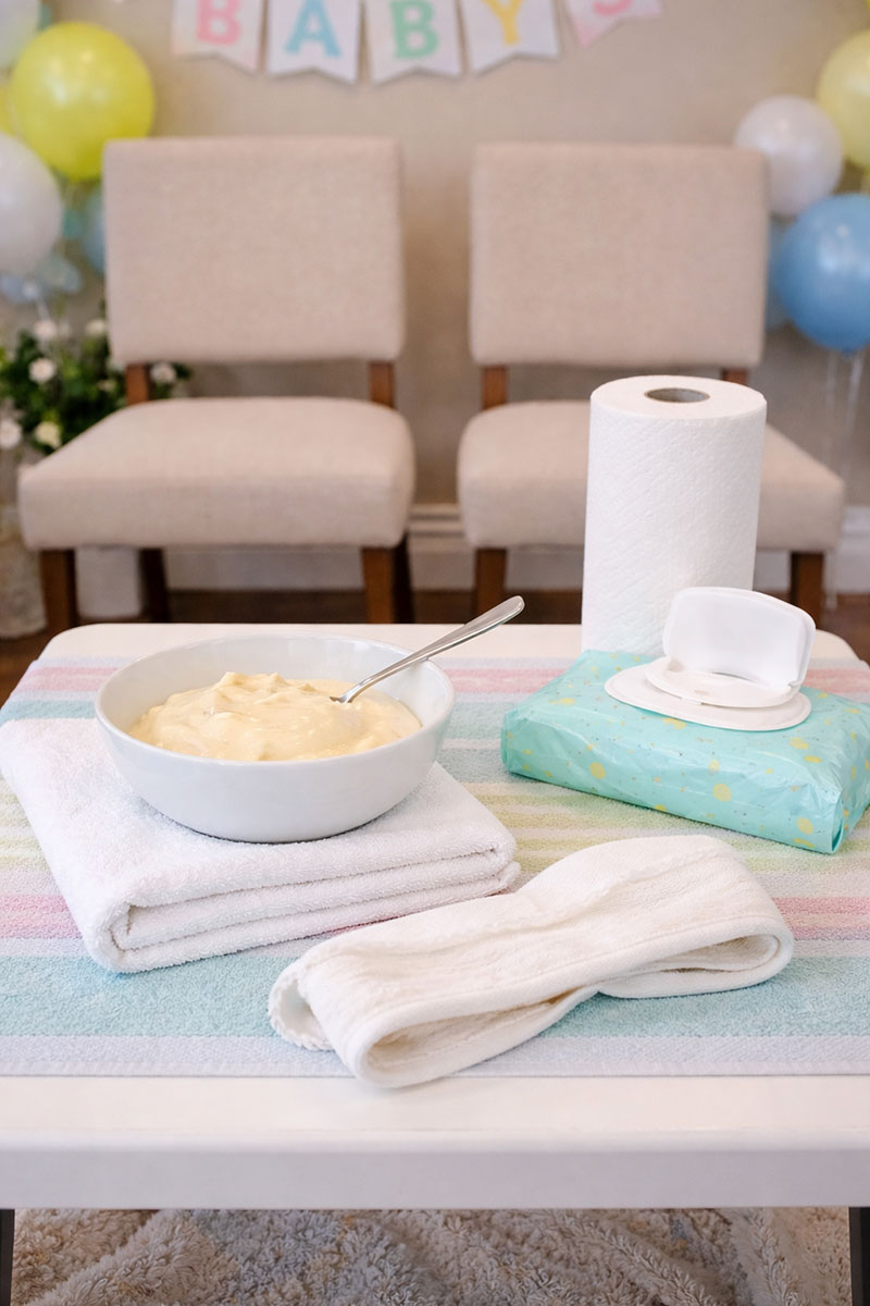 Baby shower game setup with chairs, blindfold, spoon, pudding, and cleanup towels