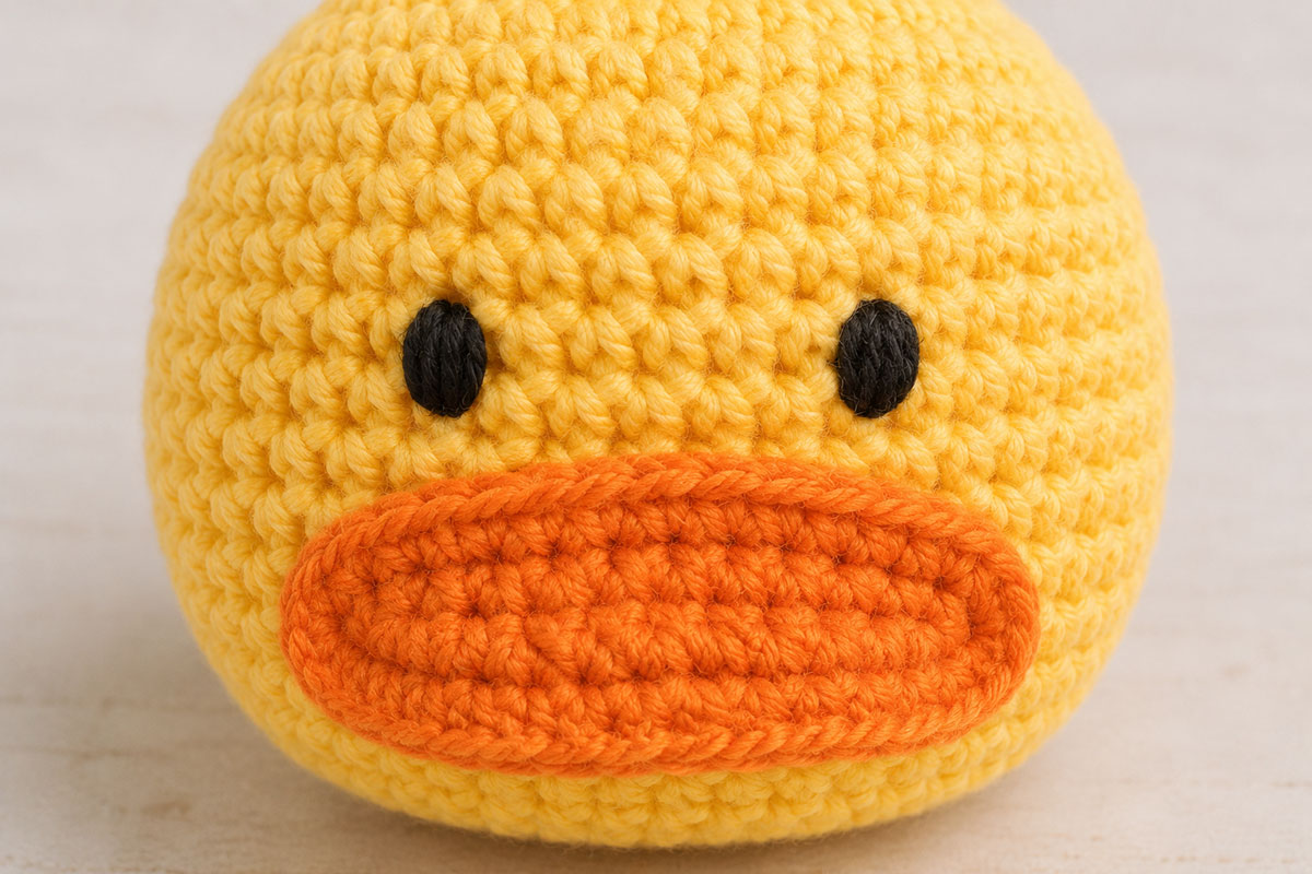 Close view of the shaped orange crochet duck bill centered beneath two black embroidered satin stitch eyes