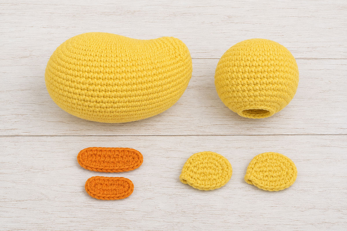 Crochet rubber duck body, head, two orange bill pieces and two yellow wings arranged before assembly
