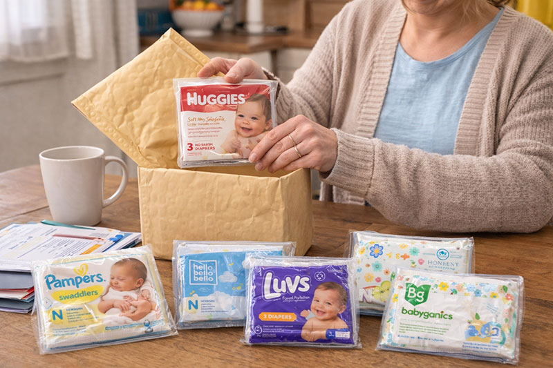 Woman opening mail at a kitchen table with several diaper sample packs laid out