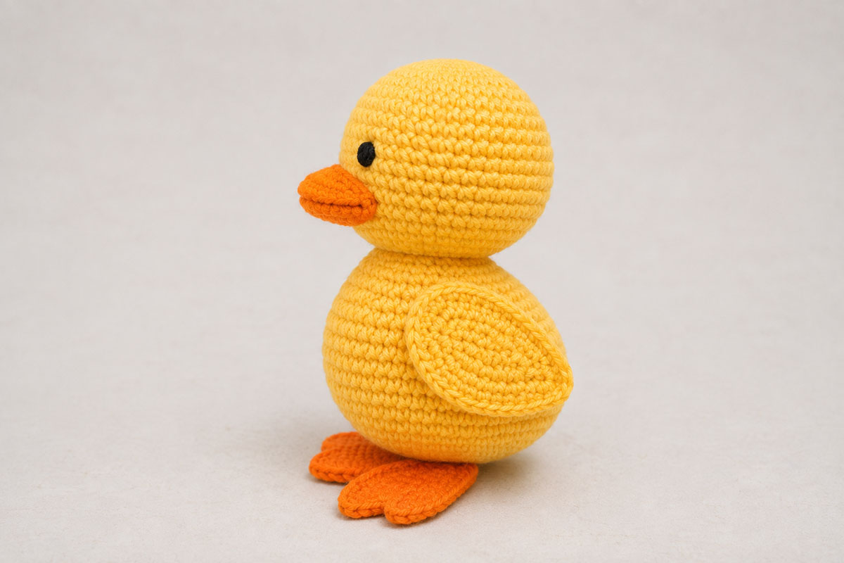 Side view of the finished yellow crochet duck standing upright with a rounded body, flat wing, orange bill and webbed feet