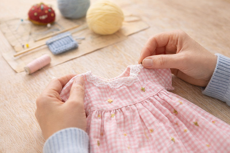 Hands sewing a small handmade baby dress using a simple sewing pattern with yarn and tools nearby