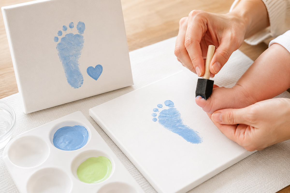 Washable children's paint, foam brushes and finished baby handprint and footprint keepsakes arranged on a protected craft table