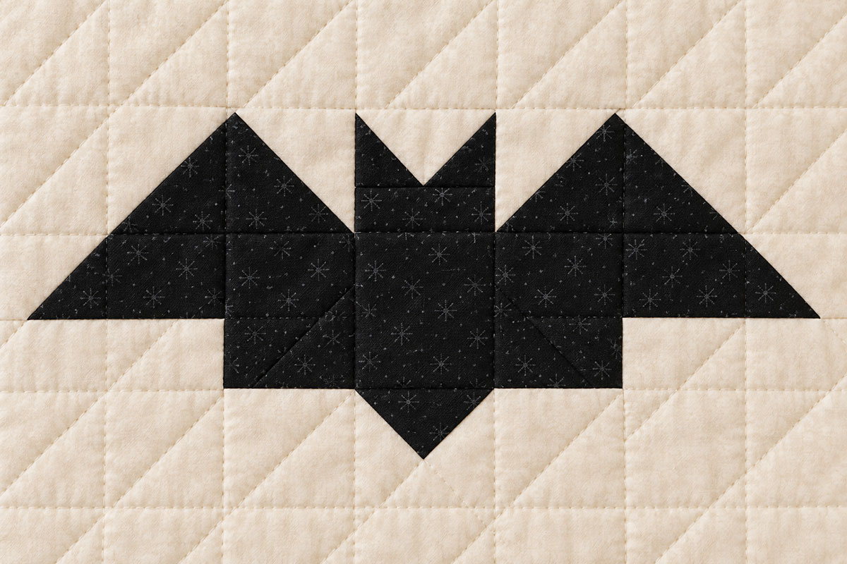 Close view of a rotary cut black patchwork bat block with two pointed ears and angular wings on a cream background