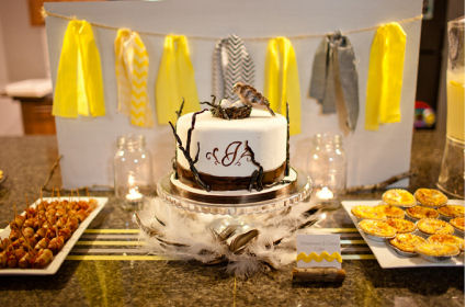 A fondant covered baby shower cake with a bird's nest and eggs cake topper surrounded by savory food