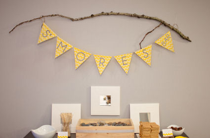 Smores bar at a baby shower with yellow chevron stripes banner displayed on a rustic tree branch