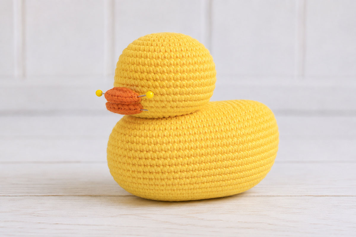 Yellow crochet duck with round head sewn to the front upper body and orange bill pieces pinned in position