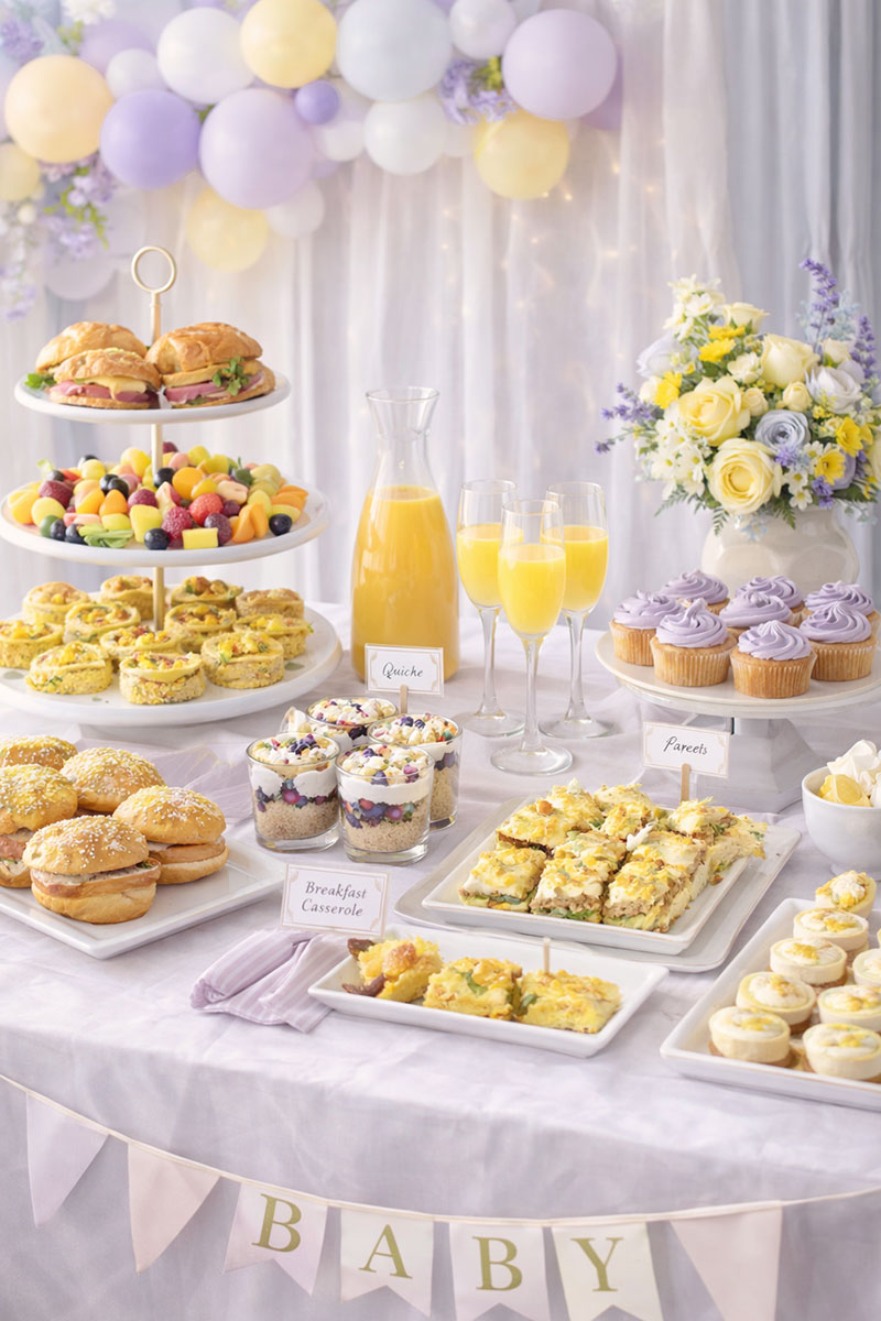 Baby shower brunch table with pastries, fruit, mini quiches, and juice in a bright magazine-style party setting