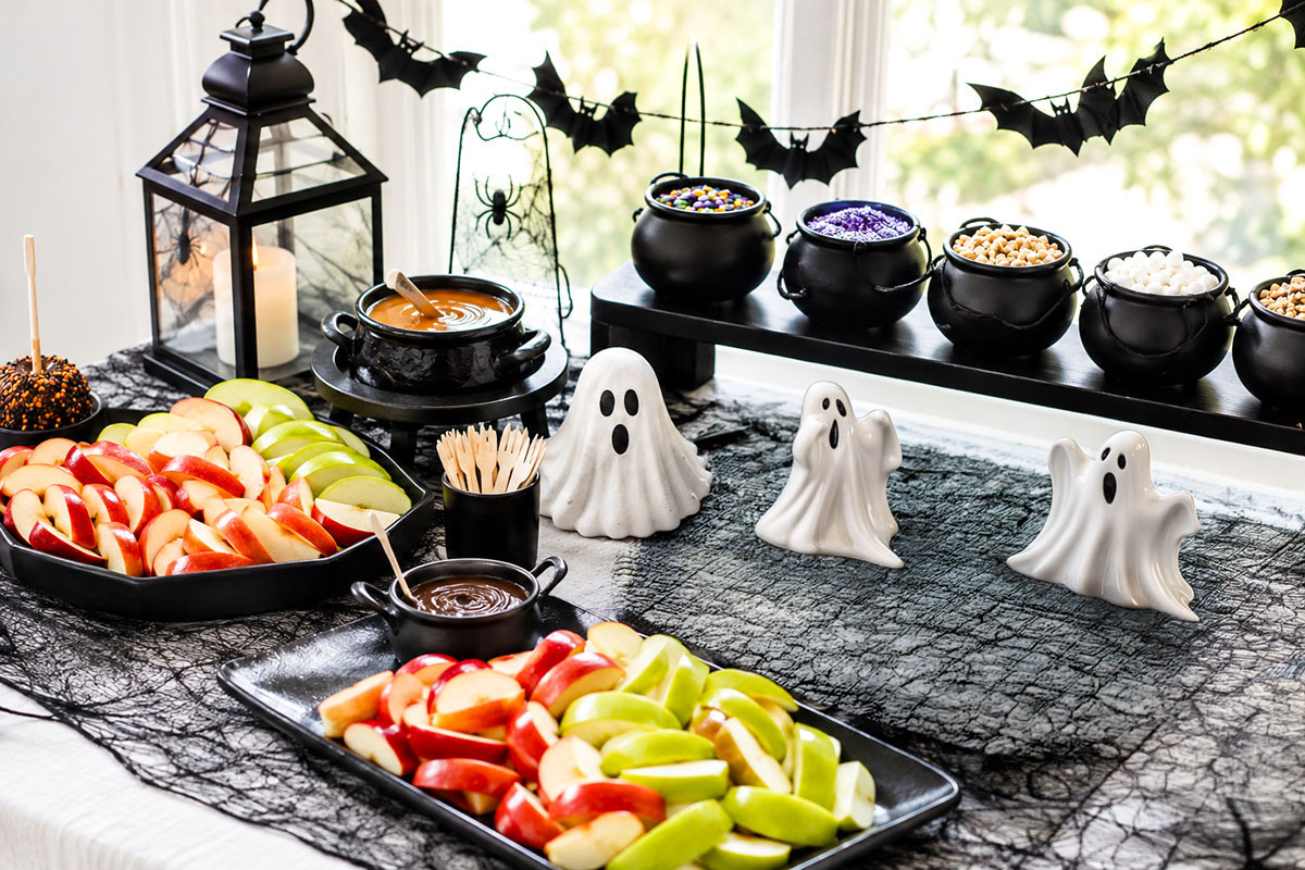 Halloween caramel apple bar with red and green apple slices, warm caramel, chocolate dip and toppings in black cauldron bowls