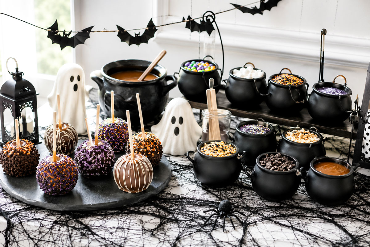 Casual creepy caramel apple bar for a Halloween baby shower with caramel apples, black cauldron topping bowls, ghost decorations and a bat garland