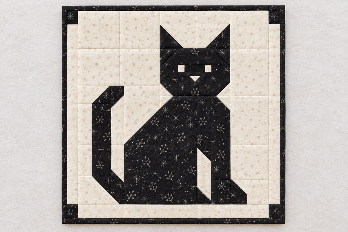 Finished pieced black cat quilt block with pointed ears, embroidered face and upright patchwork tail on a cream background