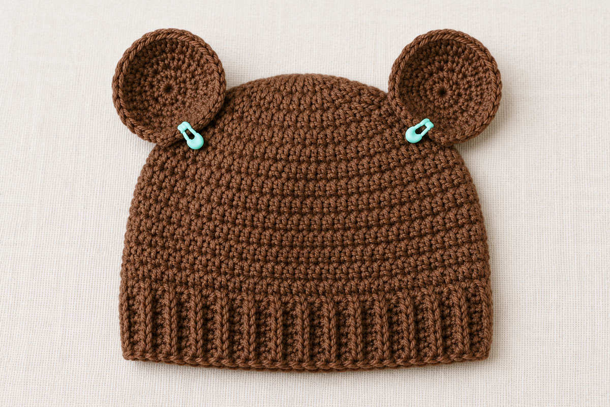 Brown crochet baby bear hat with two rounded ears held in place by stitch markers before sewing