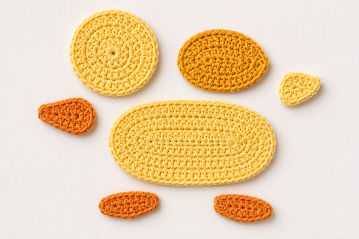 Seven flat crochet duck applique pieces with a broad yellow oval body, round yellow head, smaller gold oval wing, small yellow tail, orange beak and two orange feet