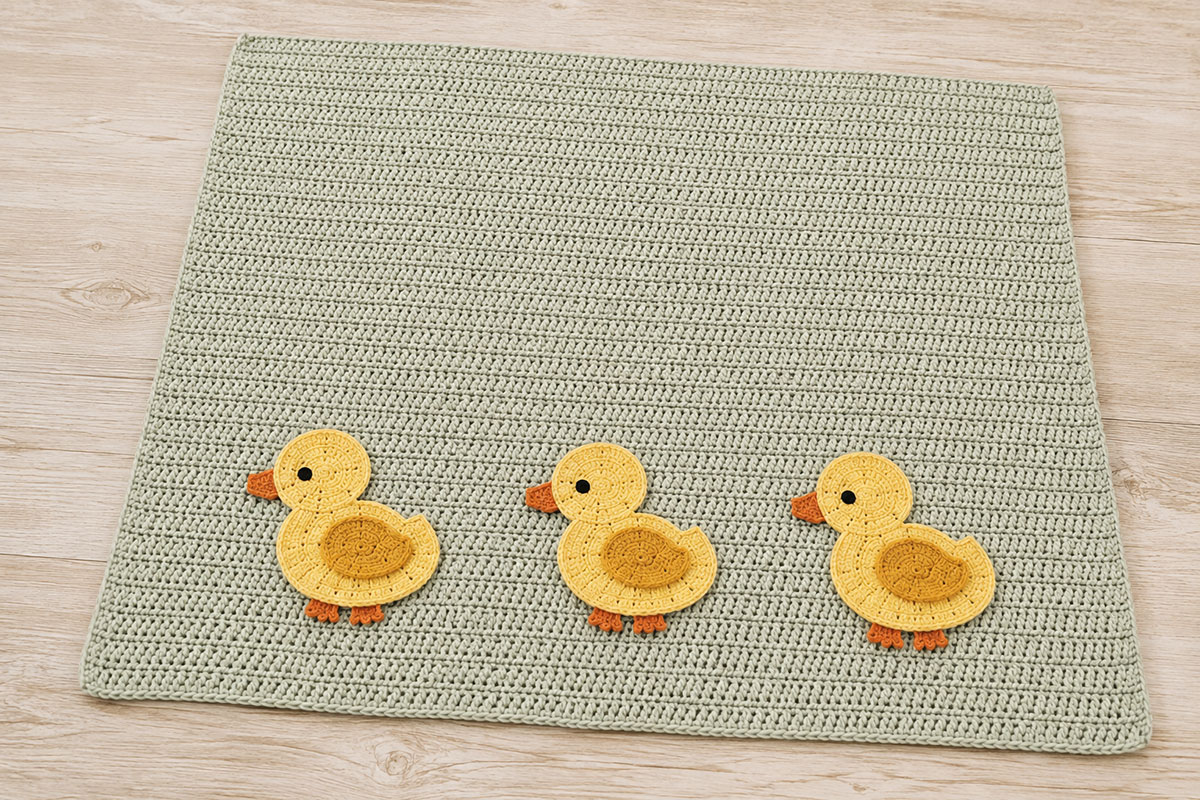 Three yellow crochet duck appliques arranged evenly in one row near the lower edge of a sage baby blanket before sewing