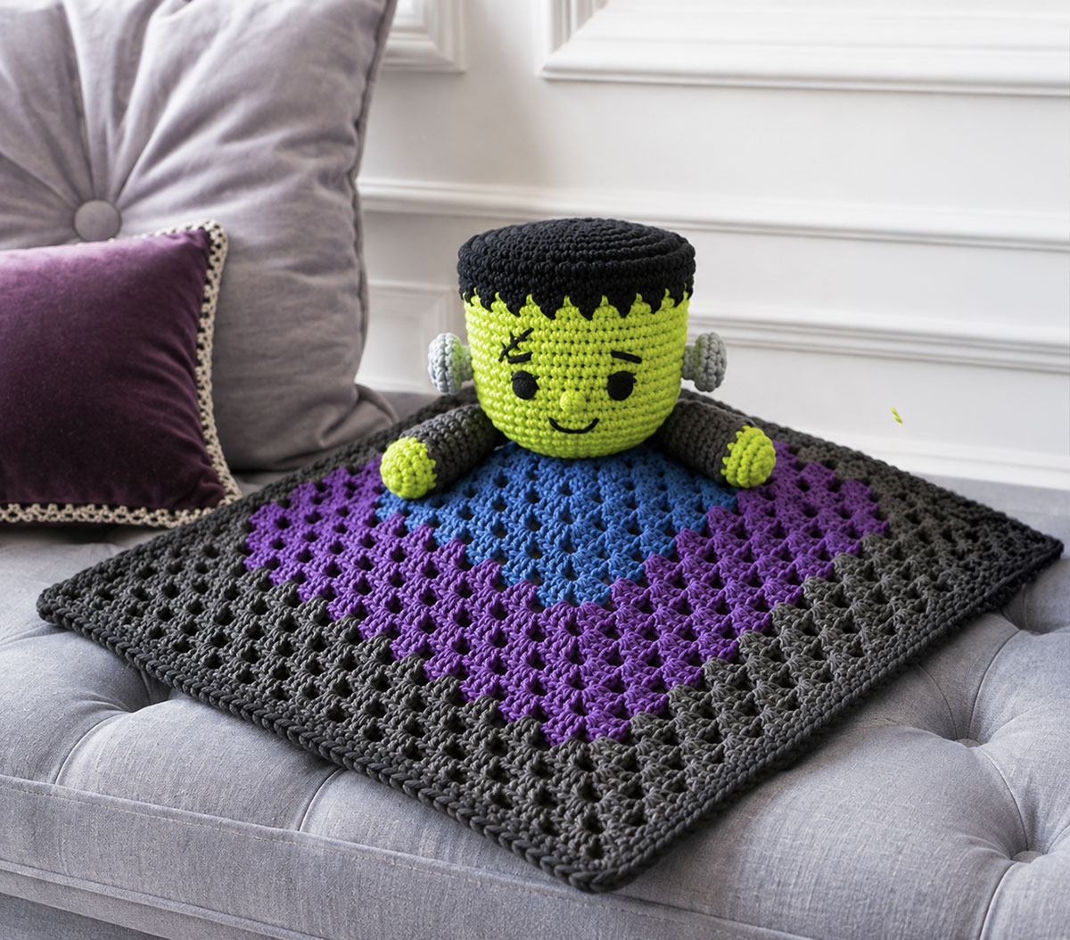 Finished crochet Frankenstein monster baby lovey with an embroidered green face black yarn hair soft gray bolts and a square blue purple and gray blanket
