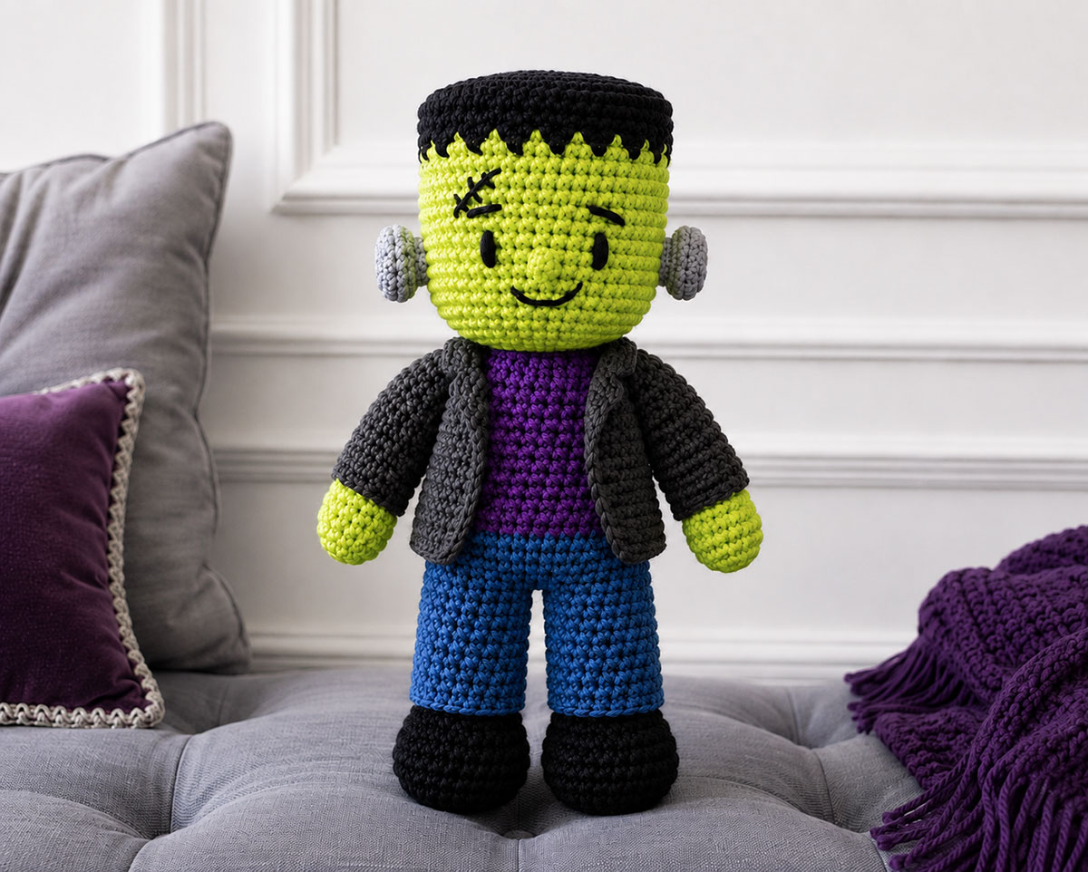 Completed crochet Frankenstein monster doll with green embroidered face black yarn hair gray jacket purple shirt and blue pants