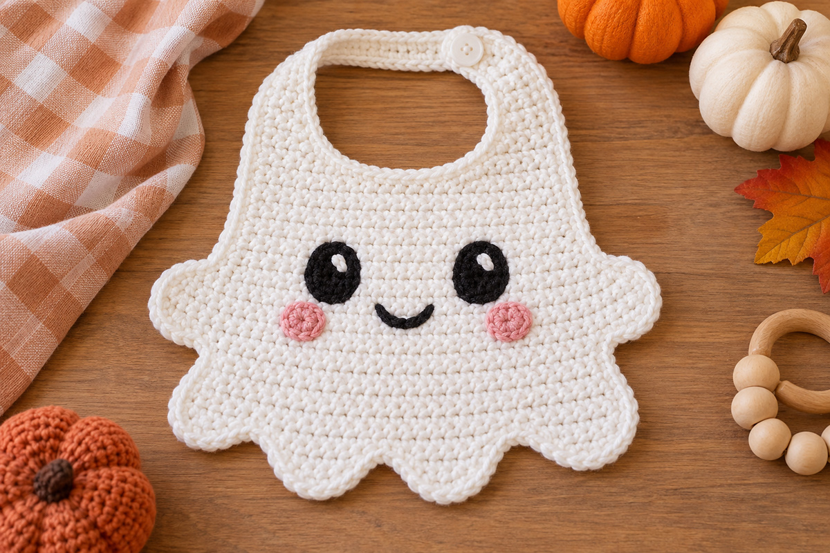 Finished white crochet ghost baby bib with rounded side arms scalloped ghost hem embroidered smiling face pink cheeks and buttoned neck strap for the free crochet ghost bib pattern