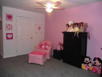 A collection of Disney Mickey and Minnie Mouse dolls and fashion dolls decorate the area to the side of the comfortable pink, upholstered nursery chair and ottoman. 