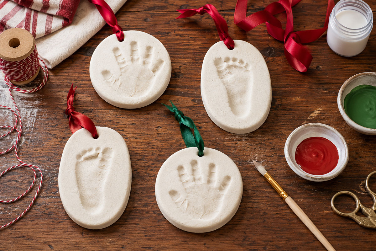 Finished salt dough baby Christmas ornaments with a handprint and footprint arranged on a wooden table with ribbon and simple craft supplies