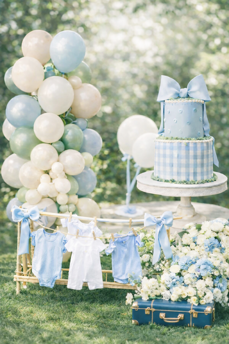 Spring and summer outdoor baby shower setup with pastel balloon garland, blue gingham cake, baby clothesline display, and blue and white flowers